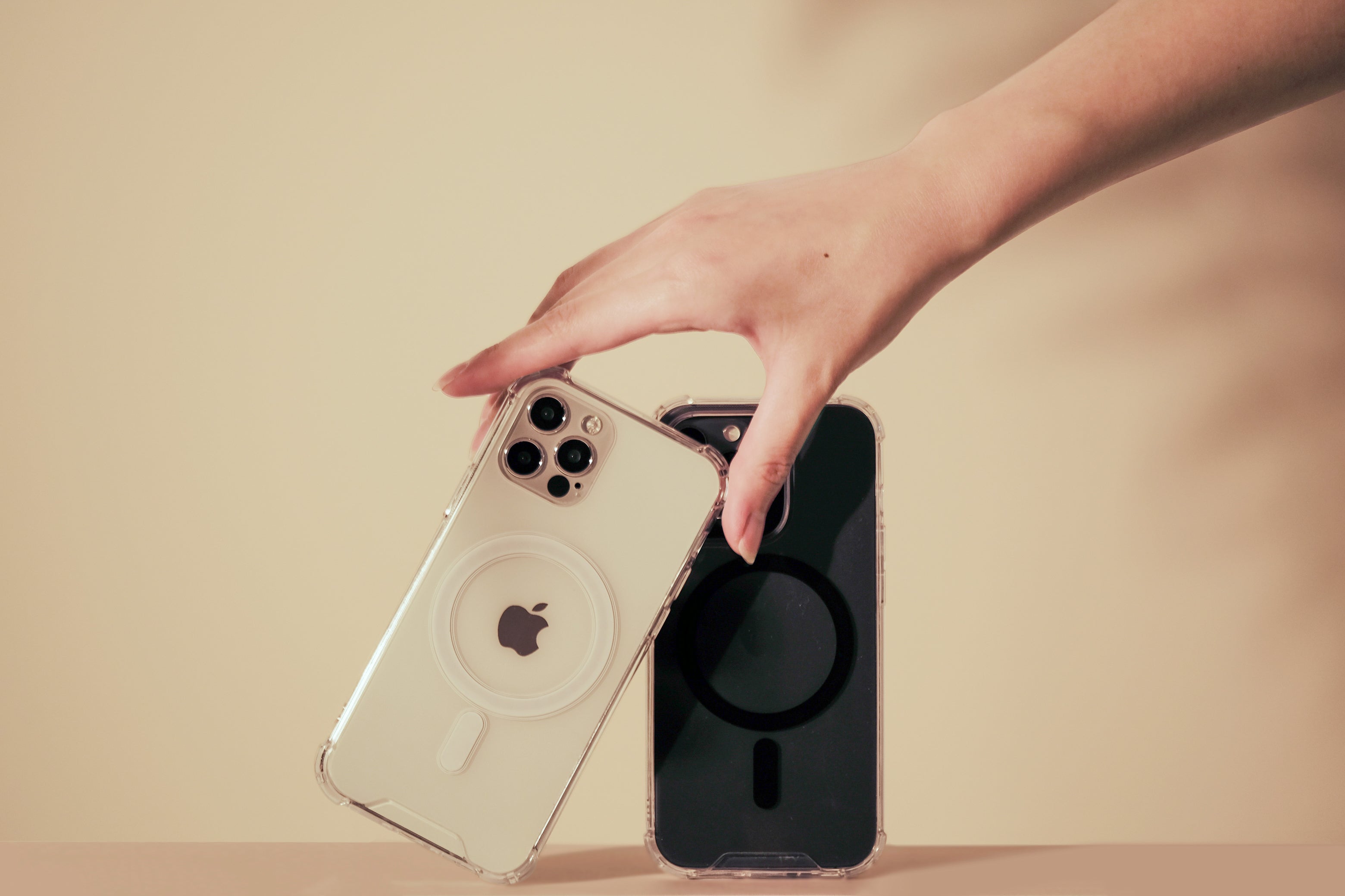 A hand holds two smartphones in clear cases. One phone is facing backward showing the Apple logo, and the other phone is facing forward with a dark screen.