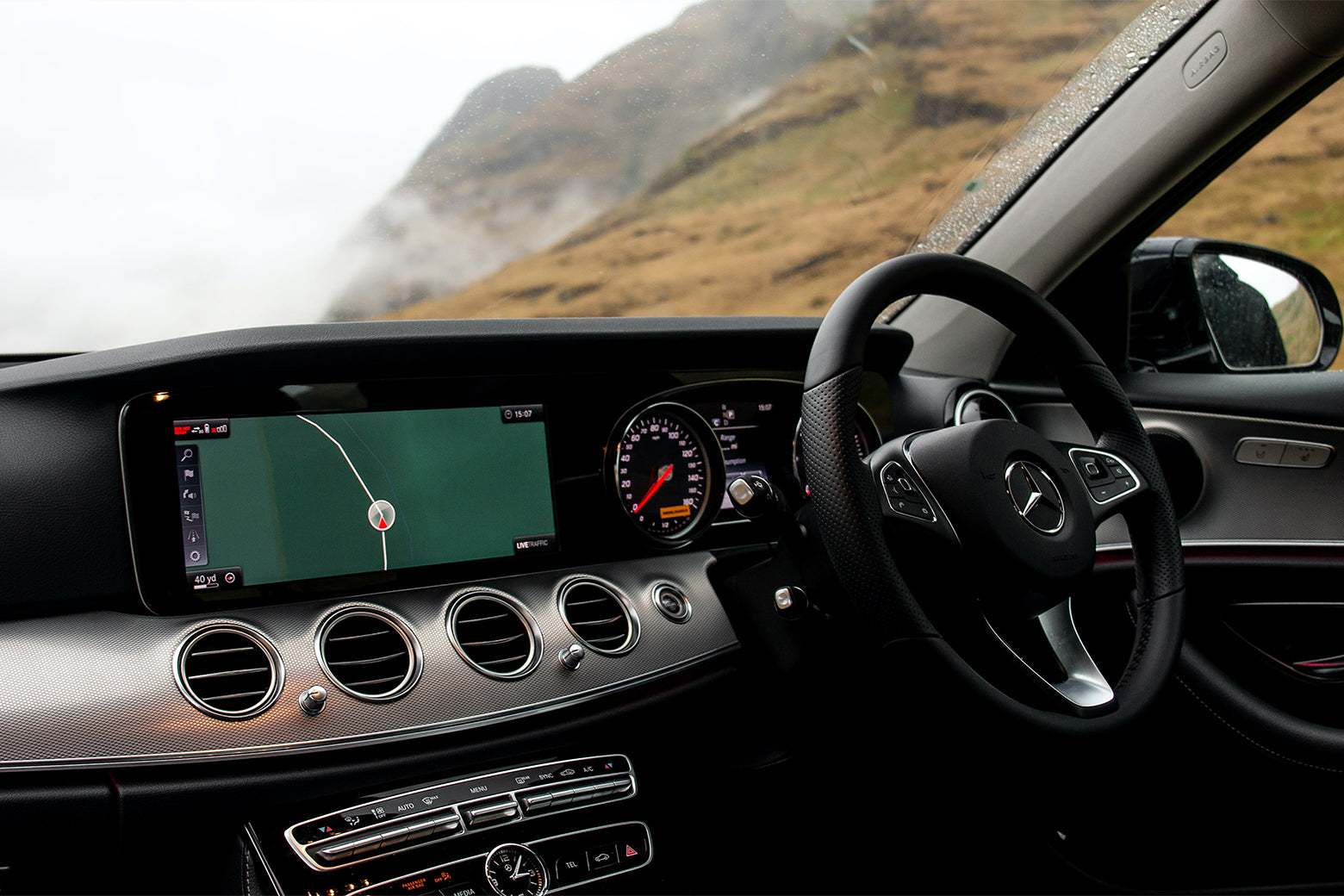 Interior of a Mercedes-Benz car showing the dashboard with a digital navigation screen, speedometer, and various control buttons. The background features a foggy landscape and hills.