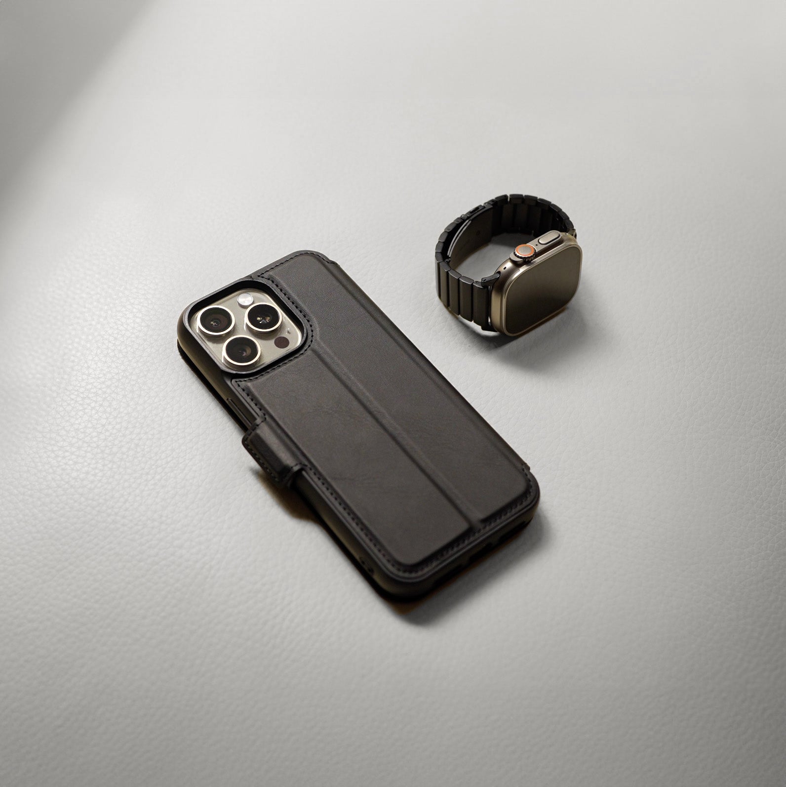 An Everyday Wallet Case for iPhone 15 Series by Urban, crafted in black leather folio, and a smartwatch with a black band are elegantly positioned on a light gray surface, emphasizing sleek protection and sophisticated style.
